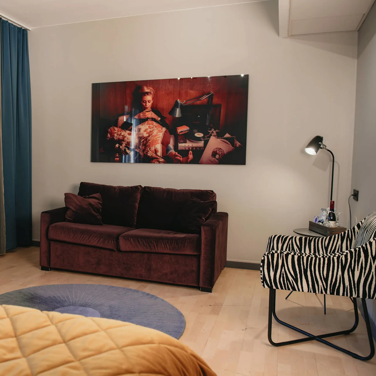 A brown sofa sits against a wall, adorned with a large photo, beside a zebra-patterned chair and a small table with a lamp, within a cozy room.