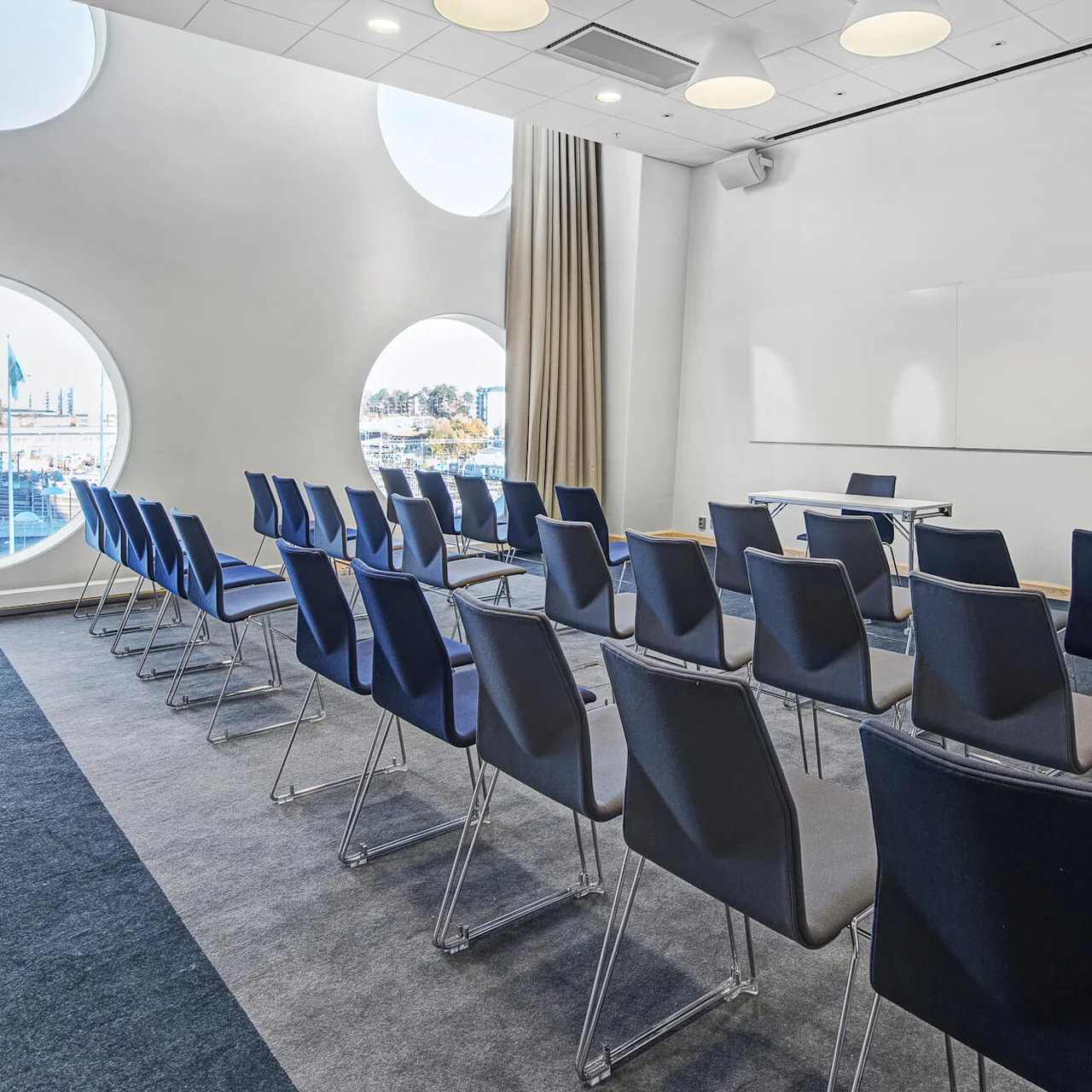 Rækker af tomme blå stole vender mod en hvid væg med en whiteboard i et moderne konferencelokale. Store cirkulære vinduer giver udsigt til en by udenfor.