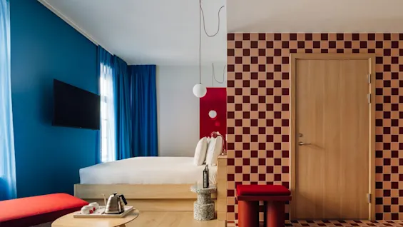 A modern hotel room with a bed, blue wall, TV, and a bold red and beige checkered pattern on the wall and floor.