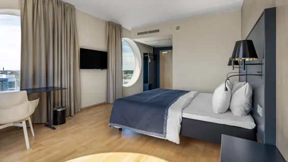 Et moderne hotelværelse med en seng, et skrivebord, et TV og et karakteristisk rundt vindue med udsigt over byen.