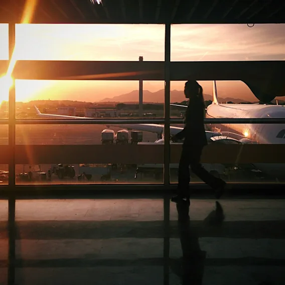 En person går genom en flygplatsterminal vid solnedgången, med ett flygplan synligt utanför fönstret.