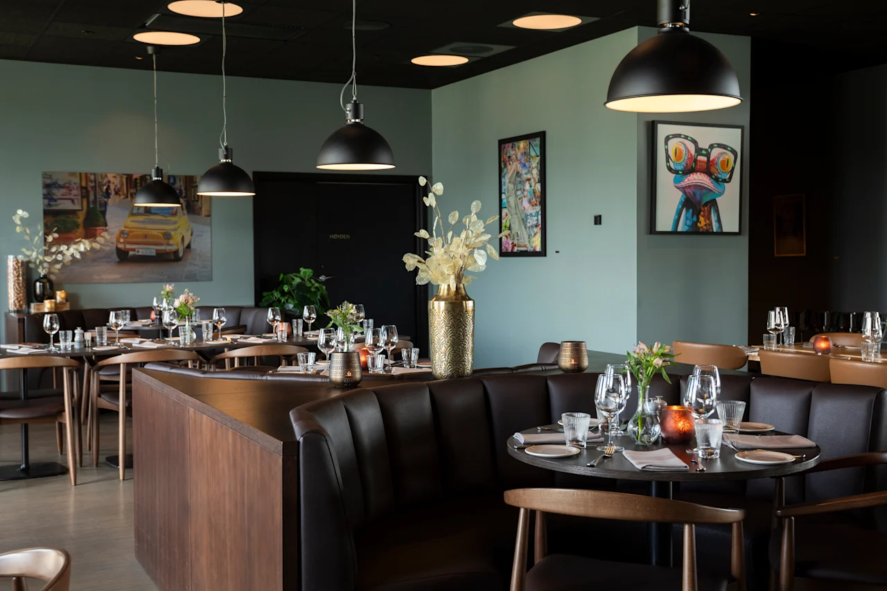 Et stilig restaurantinteriør med dekkede bord, komfortable båser og moderne kunst på veggene, som skaper en koselig spiseatmosfære.