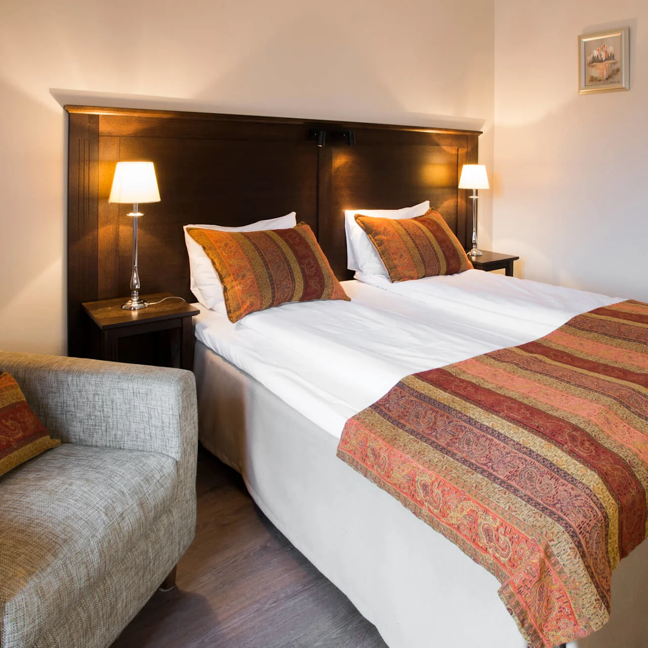 En snyggt bäddad säng med mönstrad överkast och kuddar, ackompanjerad av en stol, i ett varmt belyst hotellrum med konstverk.