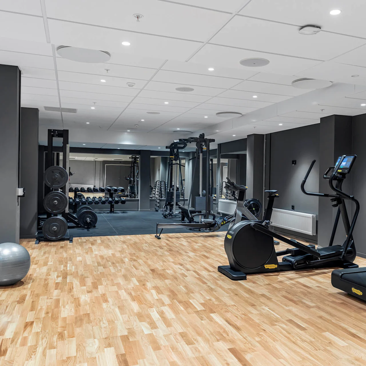 Et treningsstudio utstyrt med tredemøller og elliptiske maskiner vender mot en vegg med manualer og styrkemaskiner, alt i et romslig, godt opplyst rom med tregulv.
