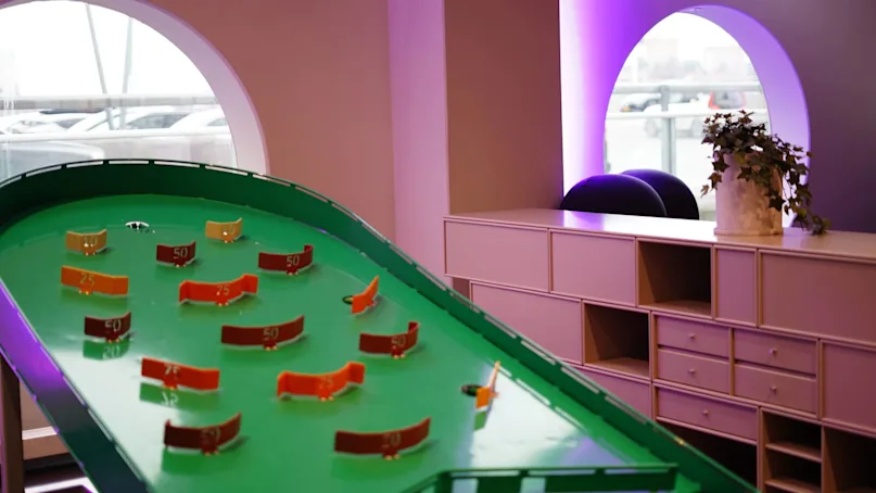 A green mini-golf game table is surrounded by point markers. It is placed in a room with a large window, drawers, and a plant, under purple ambient lighting.