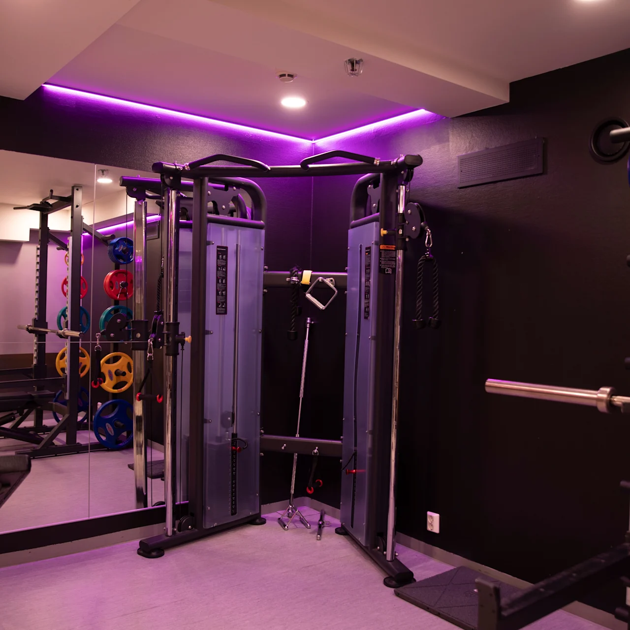 En multi-station fitnessmaskine står ubenyttet, omgivet af bænkpres og farverige vægtskiver inden for et dæmpet oplyst rum med lilla omgivelsesbelysning og spejlvægge.