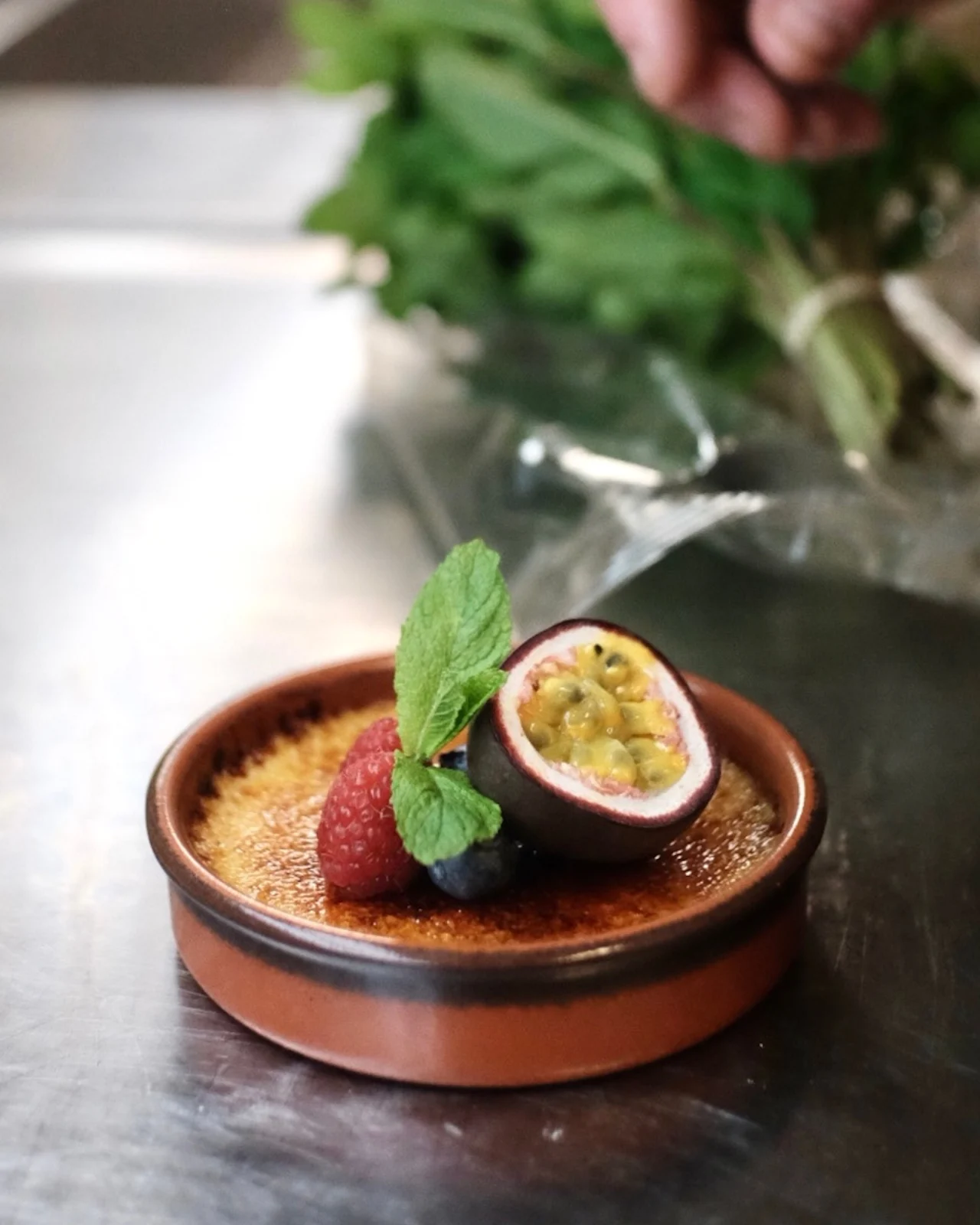 A small, round ceramic dish holds crème brûlée topped with halved passion fruit, raspberry, blueberries, and mint leaves. A hand reaches for herbs in the metallic kitchen background.