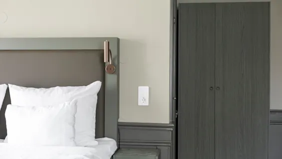 A cozy bedroom with a bed made with white linens, a wall-mounted lamp, and a dark wooden wardrobe.