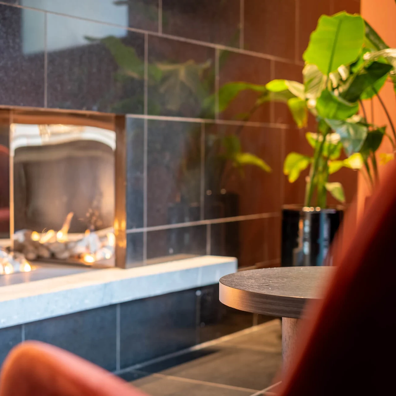 A fireplace set within dark tiled walls emits a warm flame, surrounded by a cozy interior with a plant in a pot and a round table.