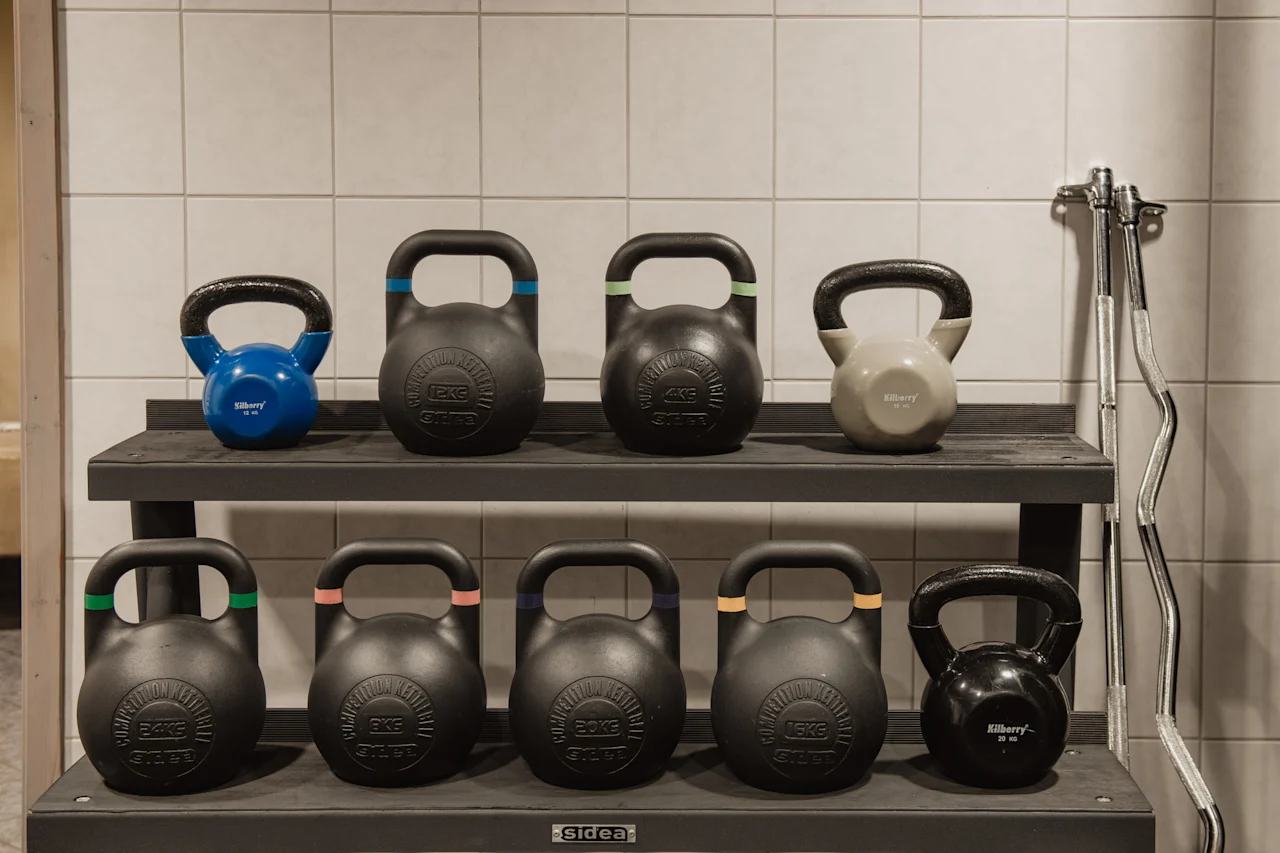 Et stativ med ulike kettlebells og en vektstang i et treningsstudio. Vektene inkluderer 8KG, 12KG, 16KG, 20KG og 24KG.