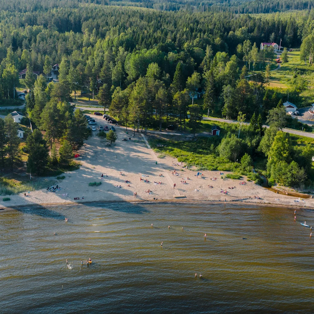 Ilmakuva vilkkaasta hiekkarannasta, jossa ihmiset uivat ja ottavat aurinkoa, vehreän metsän ja rannikkotalojen ympäröimänä.