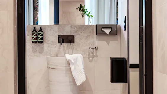 A tall, white cylindrical sink with a towel draped over it. Set against a tiled bathroom wall with a soap dispenser and tissue holder, light filters through curtained windows.