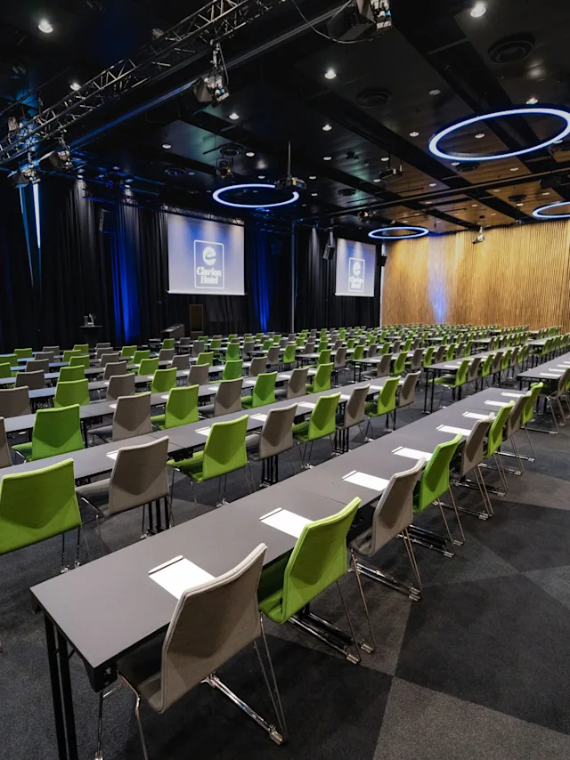 Rader med grønne og grå stoler vender mot to store skjermer som viser "Clarion Hotel", i et romslig konferanserom med geometrisk teppe og vegger av trepanel.