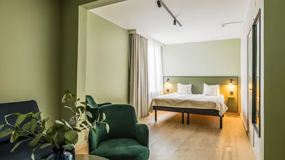 Et koselig hotellrom med en rolig atmosfære har en velredd dobbelseng mot en lysegrønn vegg, med varme leselamper. Et komfortabelt sitteområde med grønne og blå stoler tilfører eleganse.