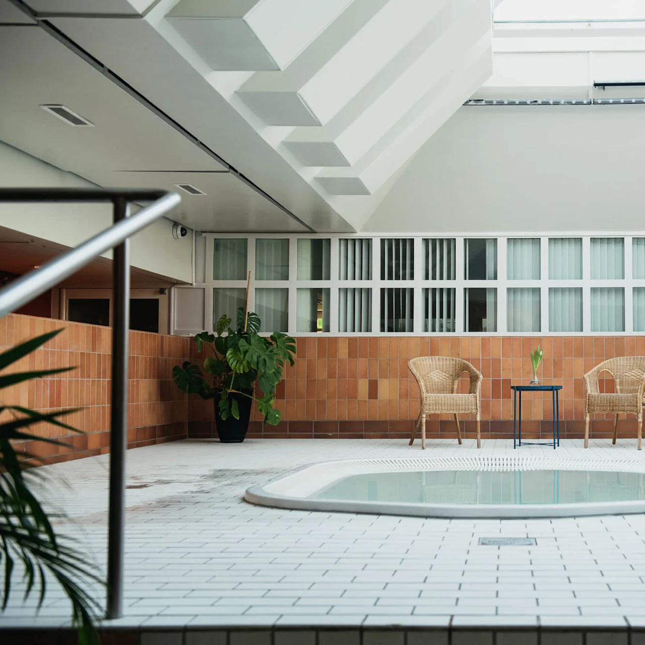 Jacuzzi with still water, surrounded by potted plants and wicker chairs on white-tiled floor, set in a bright room with large windows and brown-tiled walls.