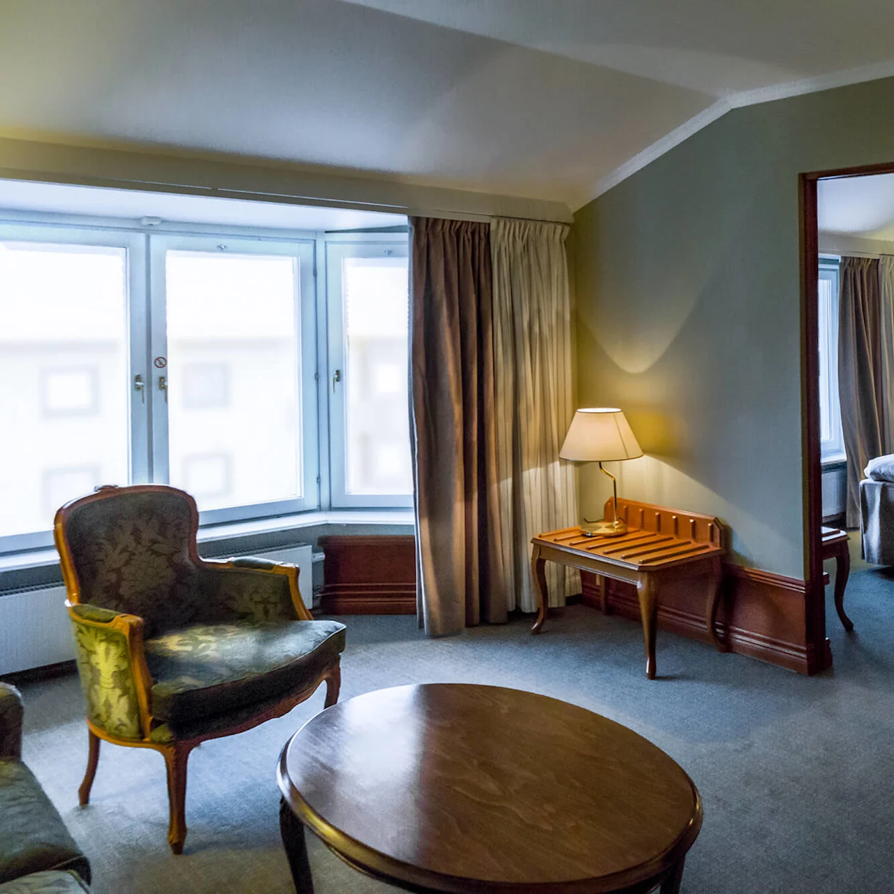 Et koselig hotellrom har en stor seng med hvitt sengetøy, nattbordslamper og en sittegruppe med lenestoler og et kaffebord ved et vindu.