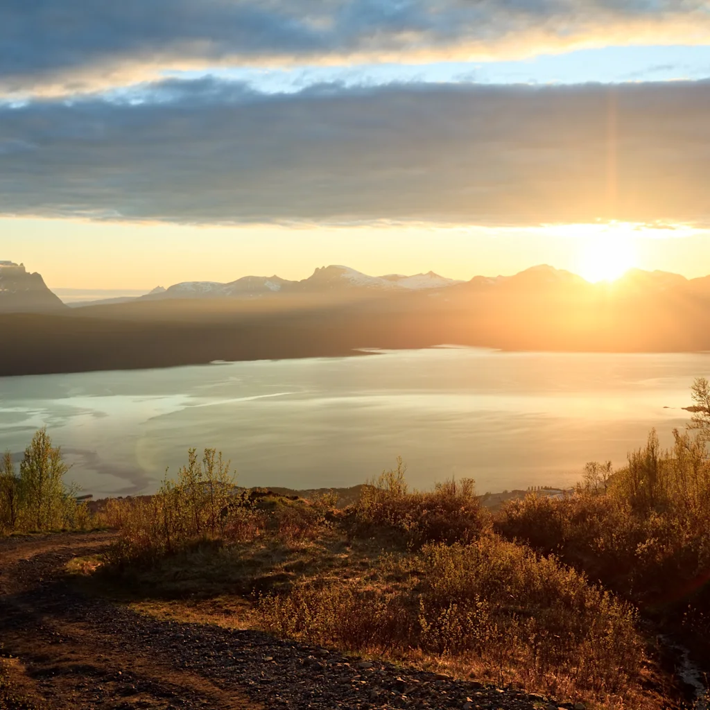 A breathtaking sunset over a vast lake, surrounded by majestic mountains and golden-hued foliage. A winding path leads into the scene.