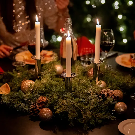 Et festlig middagsbords midtpunkt med tente lys i en grankrans med pynt, omgitt av tallerkener, glass og spisende mennesker i en varmt opplyst, juleomgivelse.
