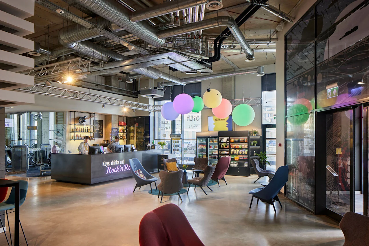 Reception desk staffed by two people, surrounded by colorful chairs and large hanging balloons in a modern lounge area. Text on the desk reads: "Keys, drinks and Rock’n’Ro."