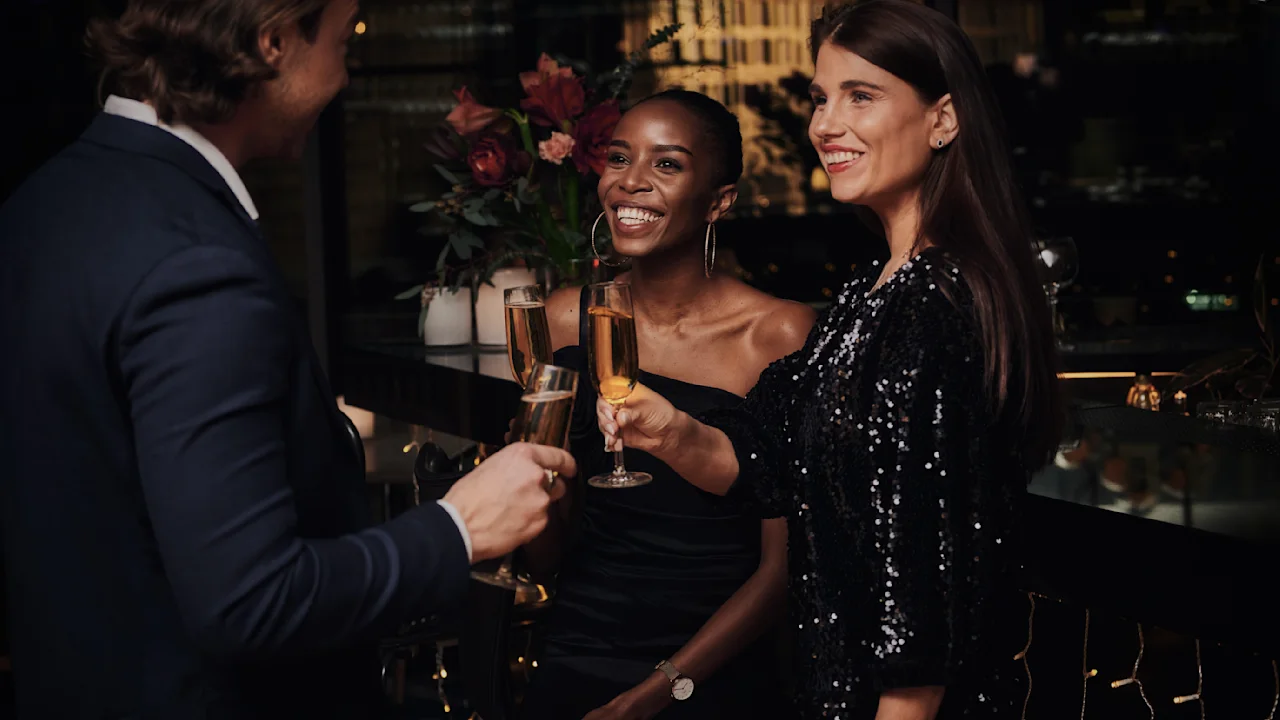 Three people in formal attire toast with champagne glasses, smiling warmly in a dimly lit, elegant venue decorated with flowers and soft lighting.