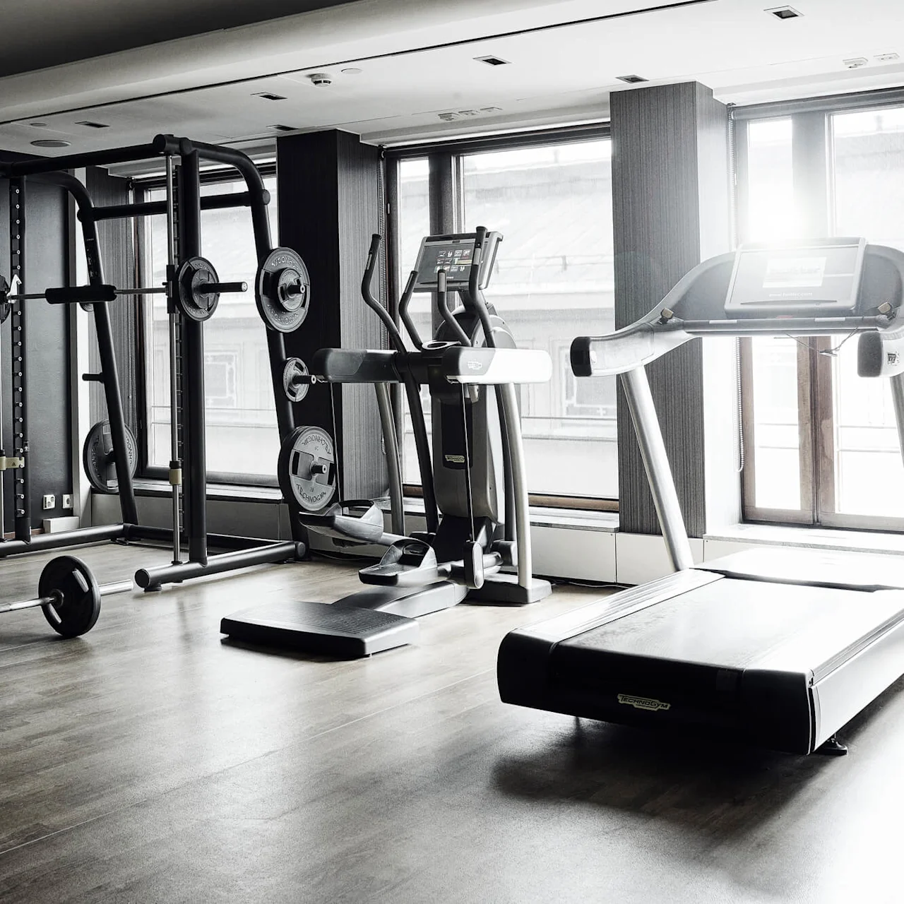 A well-equipped, unoccupied gym with various exercise machines and weights on a wooden floor, illuminated by natural light from windows.