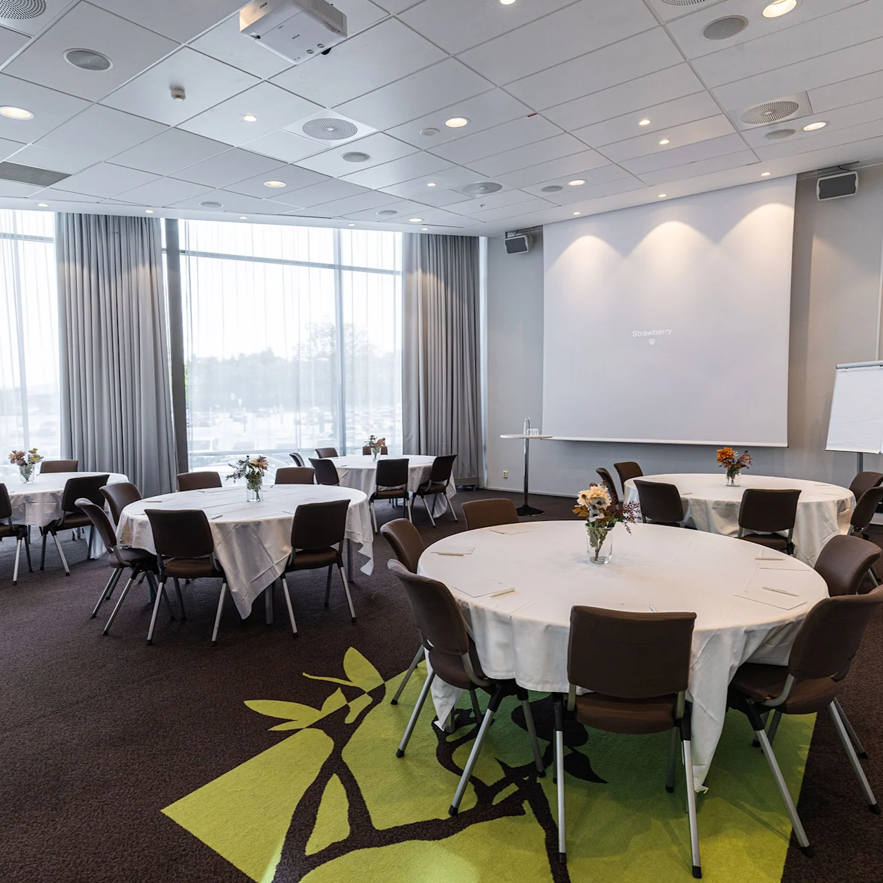 Conference room with round tables set with chairs, adorned with floral centerpieces. A projector screen, flipchart, and large windows with curtains provide a bright, professional setting.