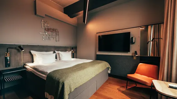 A modern hotel room with a comfortable bed, a wall-mounted TV, and a stylish orange chair, illuminated by warm lighting.