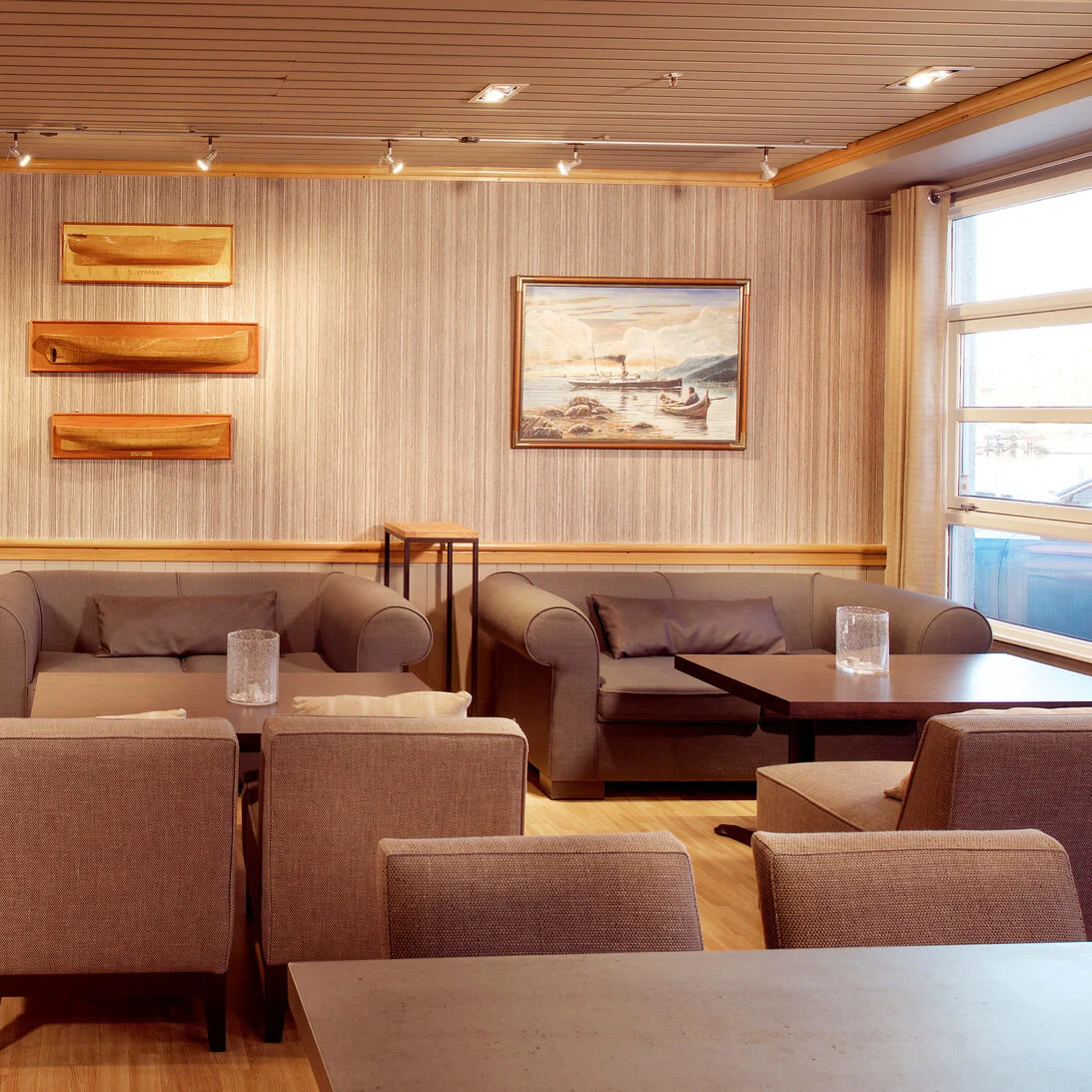 Elegant lounge med beige polstrede stole og sofaer, træhylder på stribet væg, indrammede malerier og blød belysning i et hyggeligt rum.