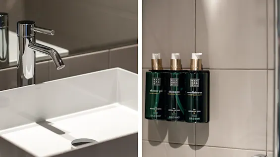 En hane over en hvid rektangulær vask med et rent, metallisk design i et badeværelse. På en flisebelagt væg er tre monterede flasker mærket "shower gel," "shampoo," og "conditioner" med "THE RITUAL OF JING."