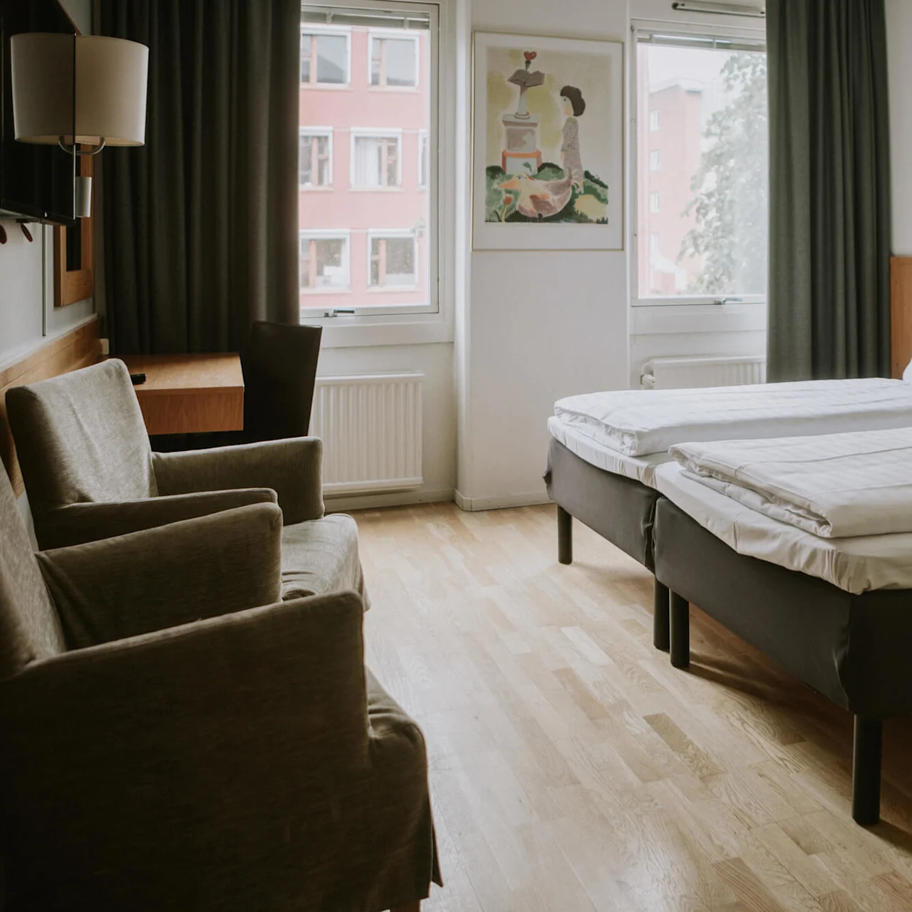 To pænt redte enkeltsenge er skubbet sammen i et hotelværelse. Stole vender mod et vindue, og et maleri hænger på væggen. Trægulve skaber en varm stemning.
