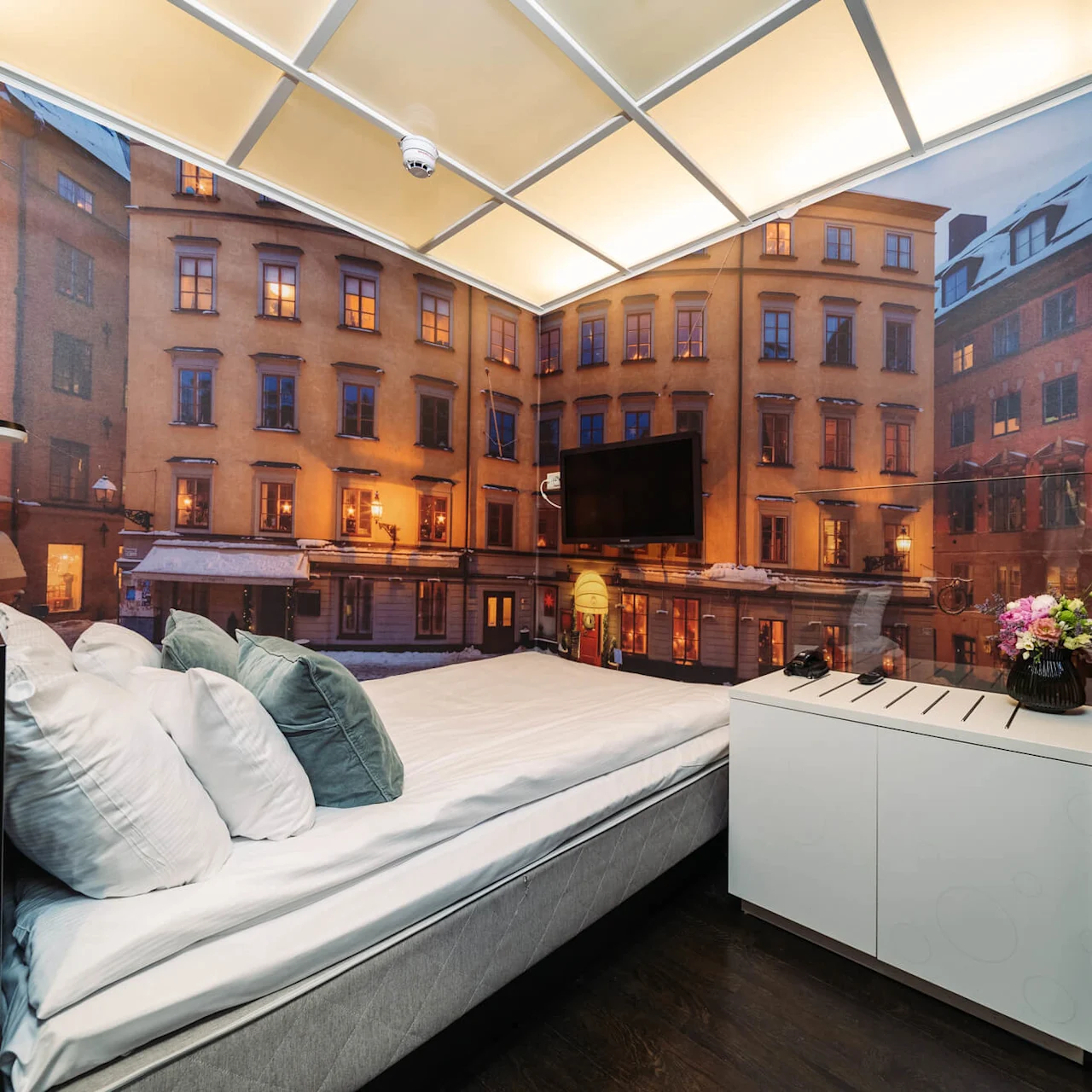 A bed with white and gray pillows sits beside a modern cabinet in a room with a mural of historic town buildings, with soft ambient lighting from the ceiling.