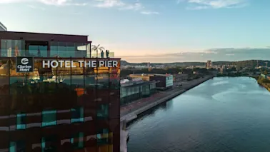 En hotelbygning med mærket "Clarion Hotel" og "HOTEL THE PIER" med udsigt over en flod. Taget har glasrækværk og er i et bylandskab under en blå himmel.