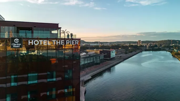 En hotelbygning med mærket "Clarion Hotel" og "HOTEL THE PIER" med udsigt over en flod. Taget har glasrækværk og er i et bylandskab under en blå himmel.