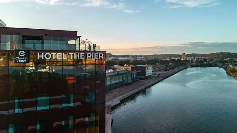 A hotel building labeled "Clarion Hotel" and "HOTEL THE PIER" overlooks a river. The rooftop features glass railings and is set in an urban landscape under a blue sky.