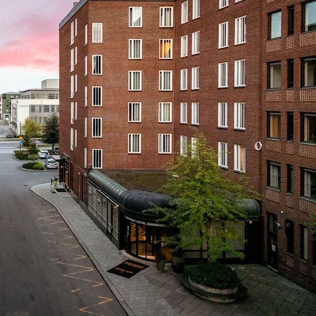 En gata kantad av höga tegelbyggnader vid solnedgång, med ett litet torg med träd och en byggnadsentré med tak som leder till en bredare väg längre bort.
