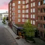 En gata kantad av höga tegelbyggnader vid solnedgång, med ett litet torg med träd och en byggnadsentré med tak som leder till en bredare väg längre bort.
