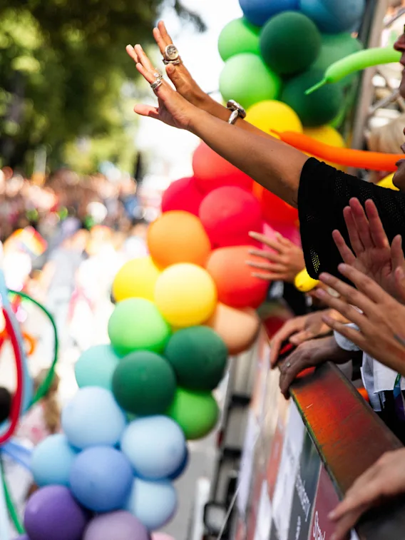 Folk jublar och vinkar från en paradvagn, med färgglada ballonger längs kanten. Den omgivande miljön är fylld av liv och rörelse, vilket antyder en festlig händelse.