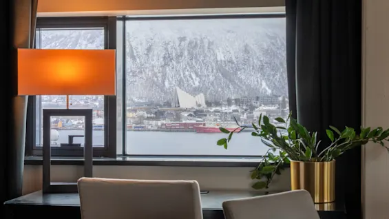 En varmt oplyst lampe står på et bord ved siden af en plante i en guldfarvet krukke, med udsigt over et snelagt bjerg gennem et vindue. Et rødt skib mærket "Tromsø Havn" ligger til kaj udenfor.