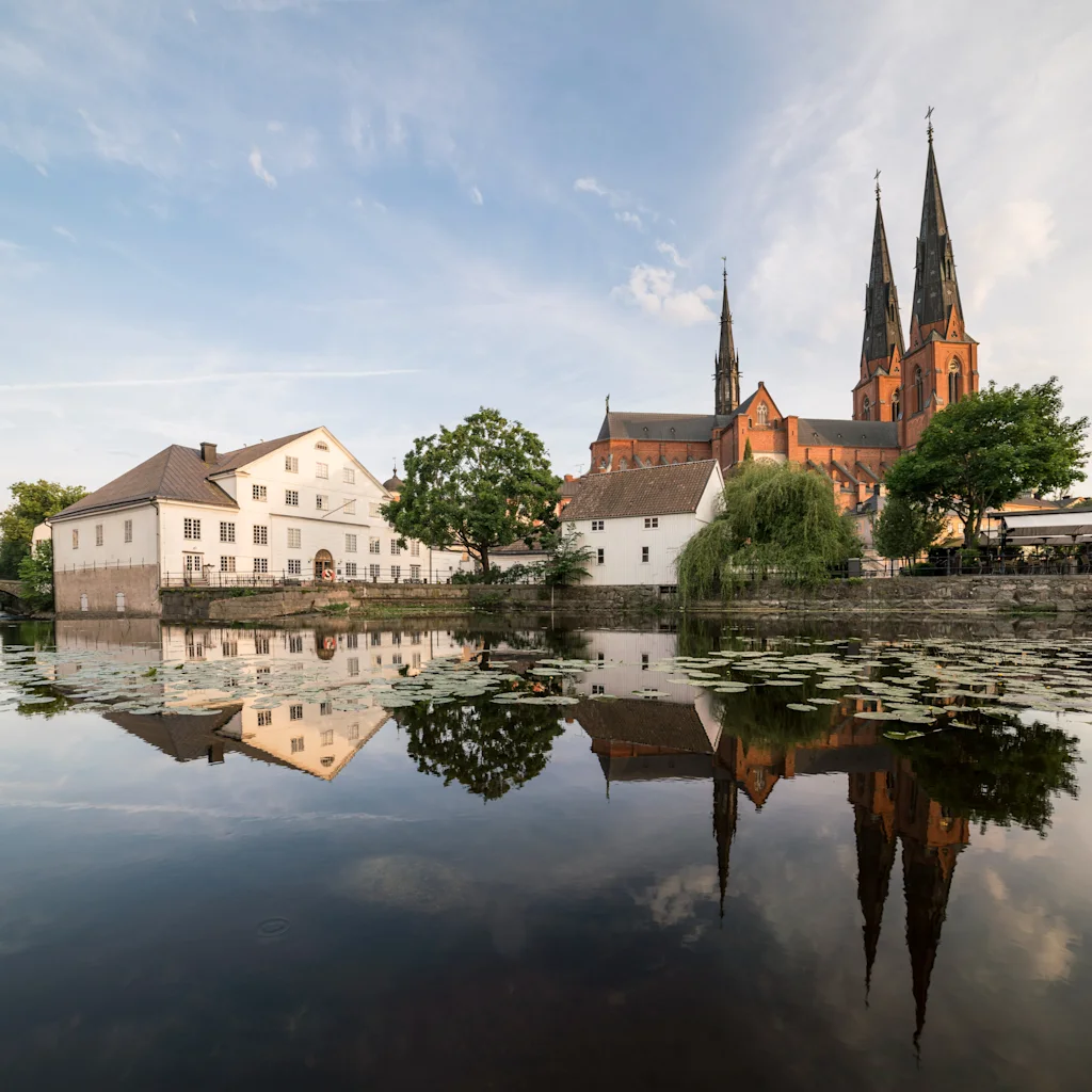 Kaunis näkymä Fyrisjokeen Uppsalassa, historialliset rakennukset ja suuri katedraali heijastuvat tyynestä vedestä.