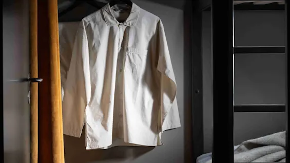 A white shirt on a hanger, attached to a wall-mounted rack, is illuminated in a dimly lit room with a bunk bed and a yellow curtain.
