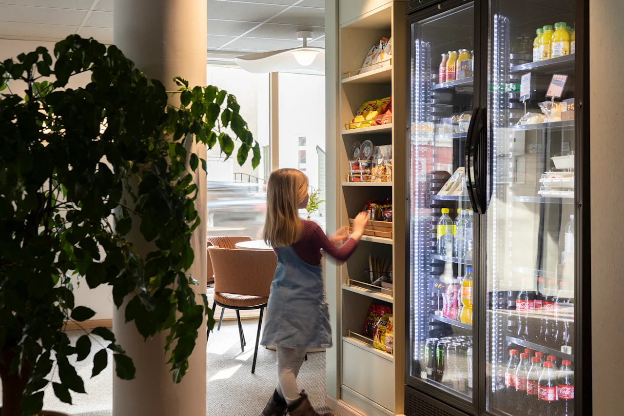 Ett barn sträcker sig efter föremål på snacks-hyllor bredvid ett kylskåp fyllt med drycker. Miljön är ett väl upplyst rum med en stor växt och stolar i bakgrunden.