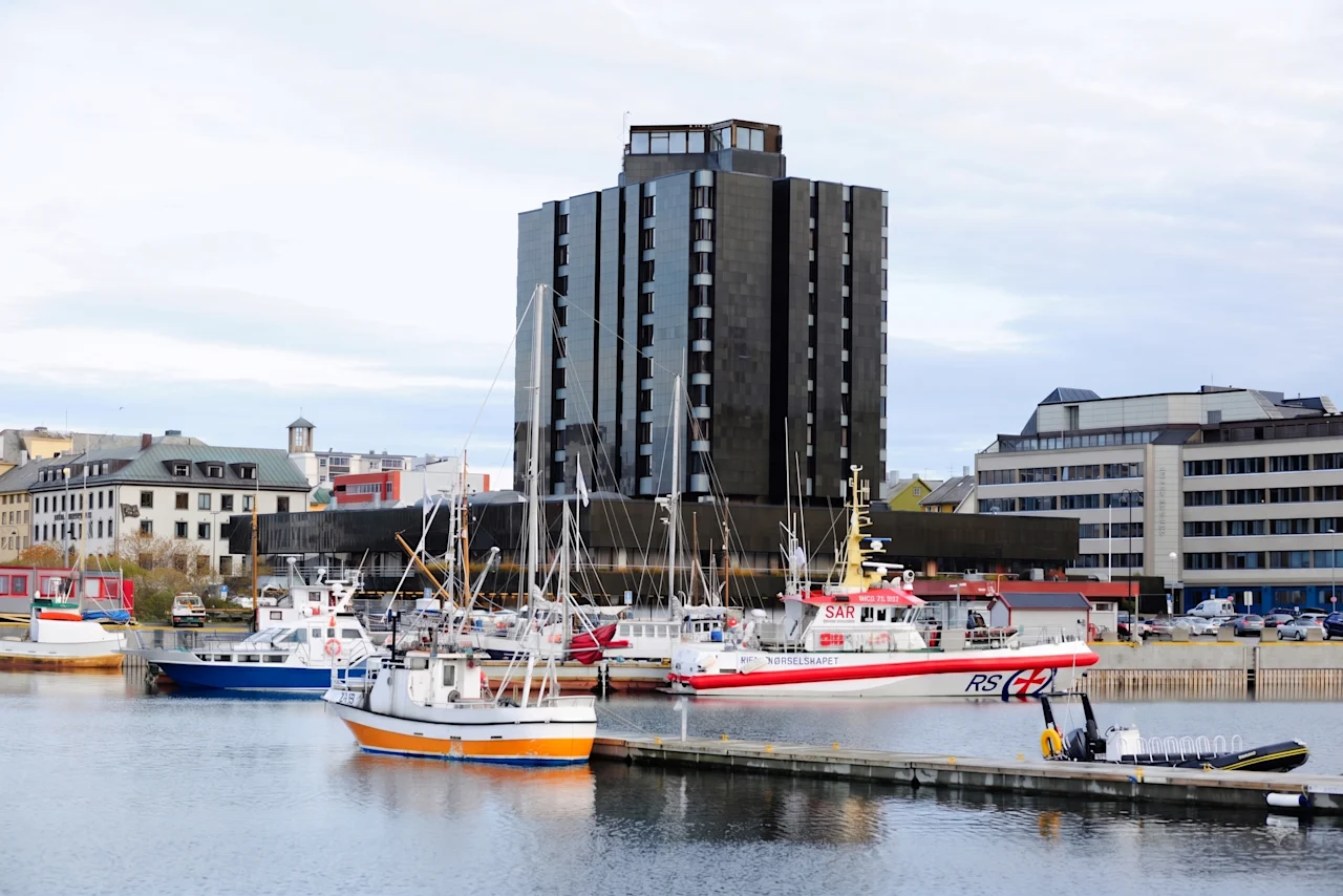 Et mørkt høybygg ved en havn fylt med båter under en overskyet himmel.