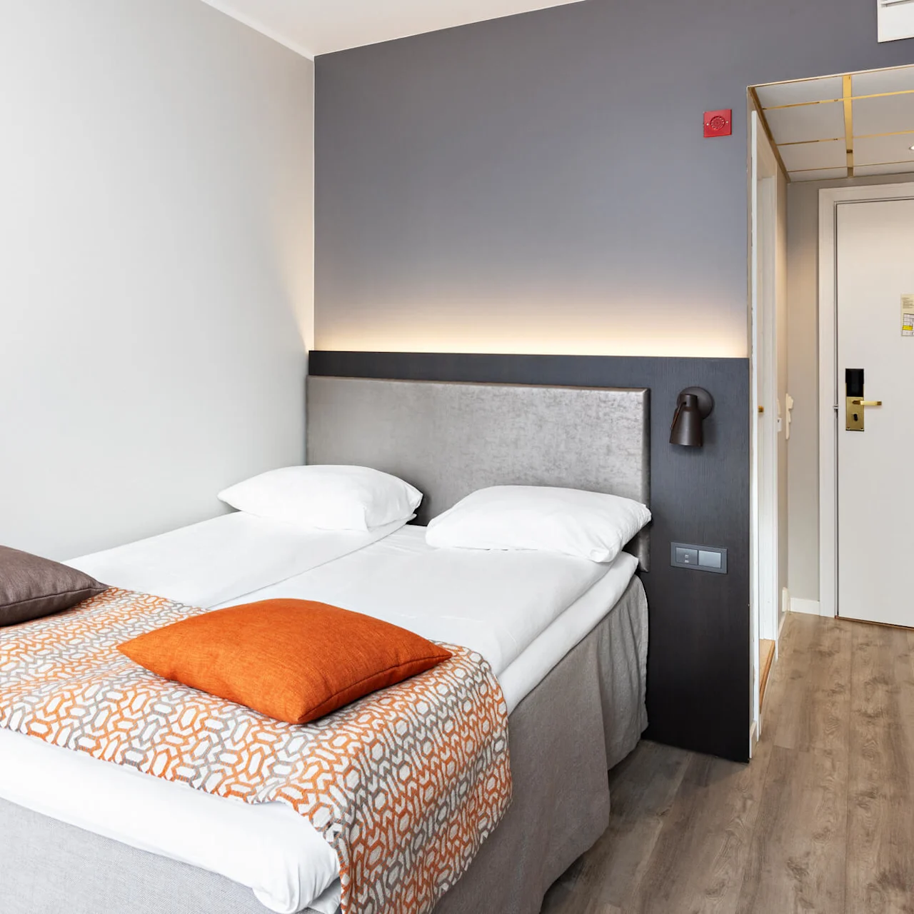 En pænt redt seng med to hvide puder, en orange-mønstret løber og farverige puder. Den står ved siden af en grå polstret sengegavl i et hotelværelse med trægulv og en lukket dør.