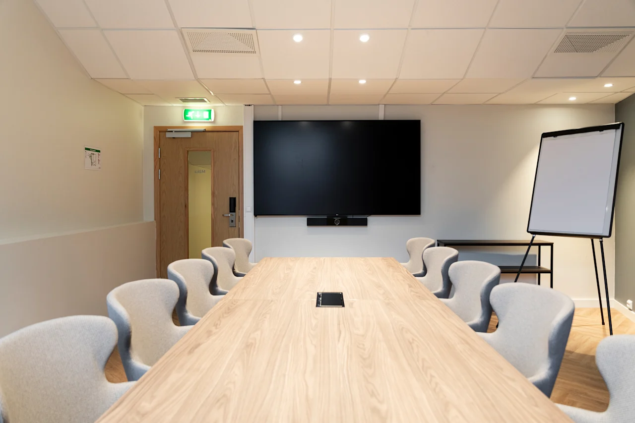 A bright and modern conference room with a long wooden table, comfortable seating, a large screen for presentations, and a whiteboard. Exit.