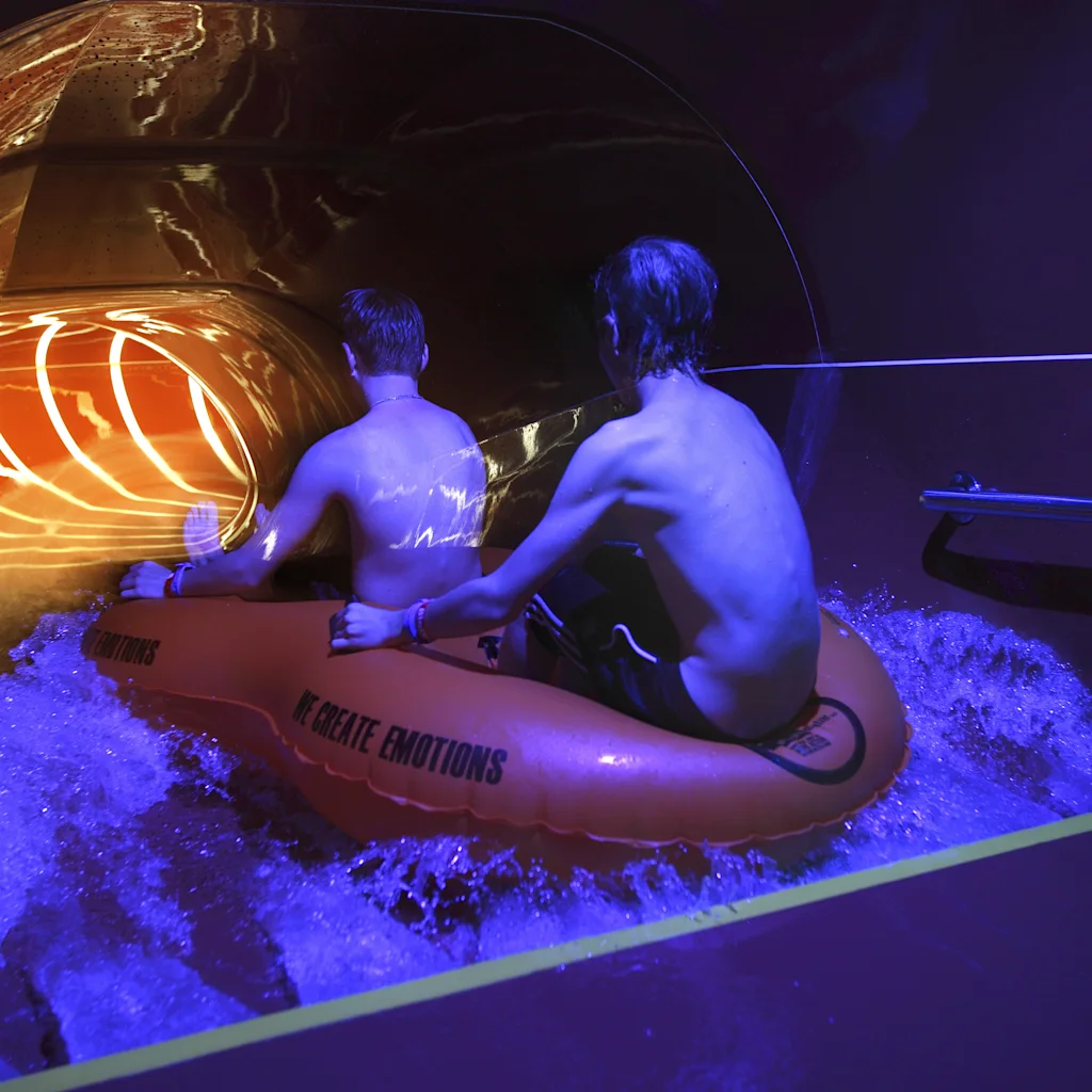 Two people on an inflatable raft exiting a brightly lit water slide at Kokpunkten Västerås, surrounded by splashing water.
