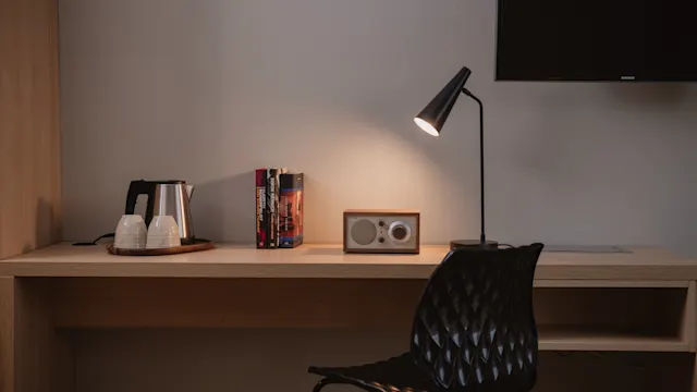 Ett skrivbord i trä med en svart stol har en lampa som lyser på böcker, en radio och en bricka med vattenkokare och koppar. En väggmonterad TV är delvis synlig ovanför.