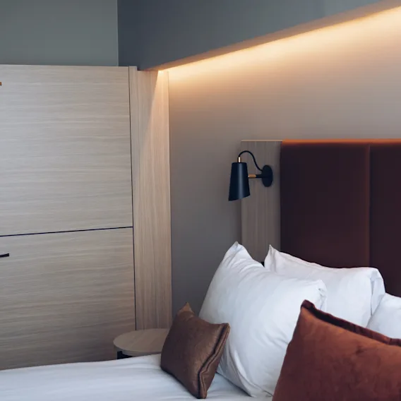 Ett modernt hotellrum med en prydligt bäddad säng, mjuka kuddar, en stilren vägglampa och varm stämningsbelysning för en avkopplande vistelse.