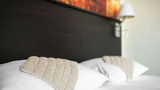 Two white pillows with quilted beige cloths lie on a bed, set against a dark wooden headboard. A wall-mounted lamp is visible in the background.