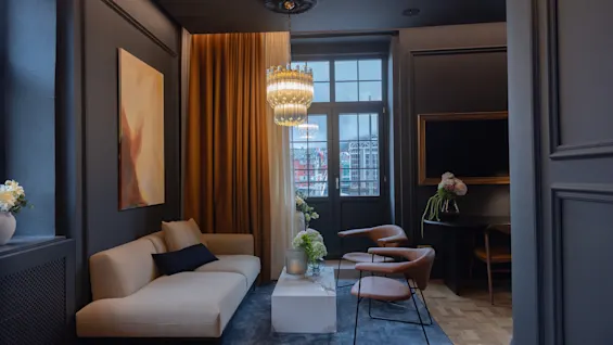 Elegant hotel suite living room with dark walls, a beige sofa, stylish armchairs, and a glowing chandelier.