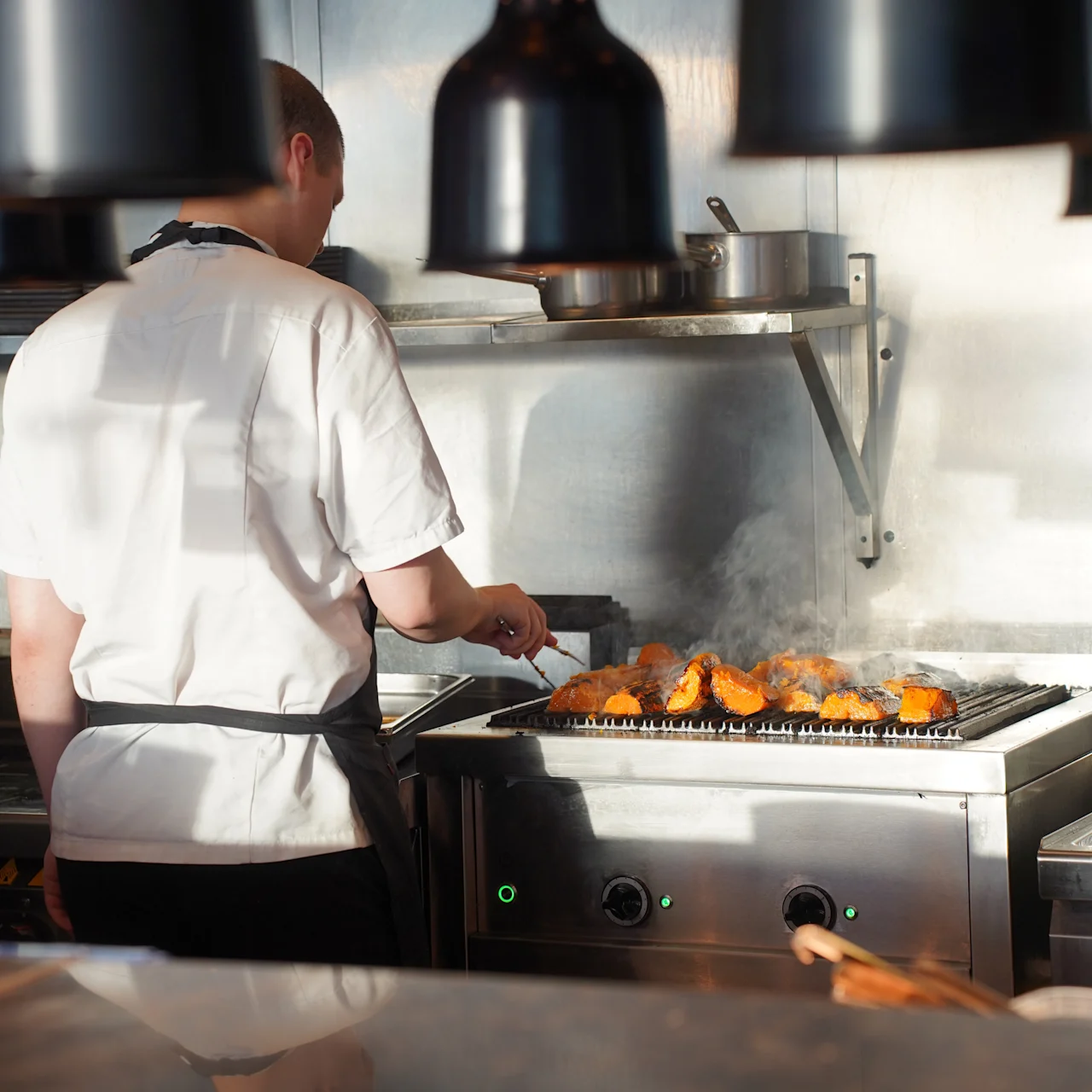 A chef grills food on a stove, surrounded by kitchen equipment and overhead lamps in a professional kitchen.